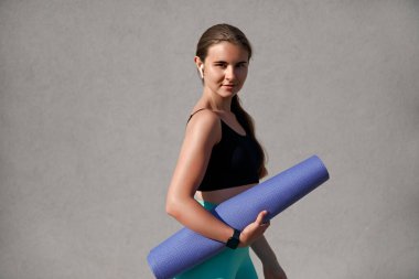 Siyah bluz ve turkuaz tayt giymiş atletik bir kadın, elinde yoga minderi tutuyor, kablosuz kulaklıklar takılı, özgüven ve sağIık yayıyor, nötr arka plan renklendirme ve zindelik üzerine kurulmuş.