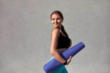 Siyah üst ve mavi tayt giymiş, elinde mor yoga minderi tutan atletik genç bir kadın, dokulu gri bir arka plana karşı özgüvenle gülümsüyor, egzersiz hazırlıklarında spor ruhu ve sağlığını somutlaştırıyor.