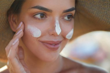 Kapalı gözlerle gülümseyen kadın yazın güneşli bir günde plajda yüzüne güneş kremi sürüyor. Cilt bakımı. Güneş koruması. 