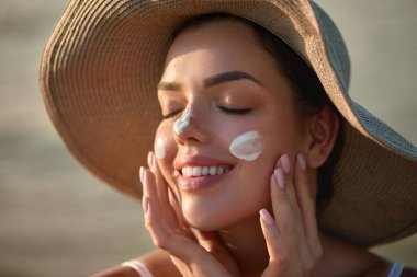 Sahilde kameraya bakarken yüzüne güneş kremi süren güzel bir kadının portresi. Cilde kadın güneş kremi sürmüş. Cilt Koruma ve Dermatoloji.