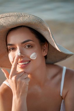 Yüzünde güneş kremi olan güzel genç bir kadın. Sahilde güneş kremi şişesi tutan bir kız. Kadın şapka cilt üzerinde nemlendirici losyon uygulayarak. Cilt bakımı. Güneş koruması. Güneş