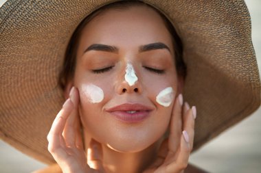 Kapalı gözlerle gülümseyen kadın yazın güneşli bir günde plajda yüzüne güneş kremi sürüyor. Cilt bakımı. Güneş koruması. 