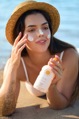 Gün batımında bikinili, gülümseyen kadın plajda güneş kremi sürüyor..