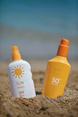 Biri beyaz diğeri turuncu olmak üzere iki güneş kremi şişesi parlak bir okyanus arka planına sahip kumlu bir sahilde sergileniyor. Güneş ve açık hava rahatlığını vurgulayan mükemmel bir yaz tatili konsepti..
