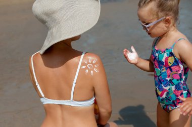 Bir çocuk, sahilde oturan bir kadına güneş kremi sürmesine yardım eder, güneş koruması ve kaliteli aile zamanı üzerine vurgu yapar, güneşli bir ortamda güneşli bir plaj oluşturur..