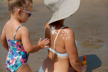 Kumlu bir plajda genç bir çocuk, güneşli gökyüzünün altında güneşli bir desen oluşturarak, renkli mayo ve rahat bir atmosferle güneş güvenliğinin ve deniz zevkinin altını çizerek bir kadına güneş kremi sürüyor..