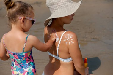 Genç bir çocuk güneşli bir sahilde oturan bir kadına güneş kremi sürmeye yardım ediyor. Görüntü, renkli bir yaz mevsiminde güneşe karşı ihtiyatlı ve özenli bir koruma sağlıyor.