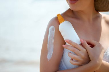 Bir kadın kumsalda güneşli bir günün tadını çıkarırken omzuna güneş kremi sürüyor. Parlak sahnede yaz etkinlikleri sırasında cilt bakımına ve güneşe önem veriliyor.