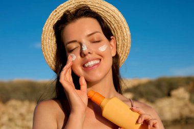 Yüzünde güneş kremi olan güzel genç bir kadın. Sahilde güneş kremi şişesi tutan bir kız. Kadın şapka cilt üzerinde nemlendirici losyon uygulayarak. Cilt bakımı. Güneş koruması. Güneş