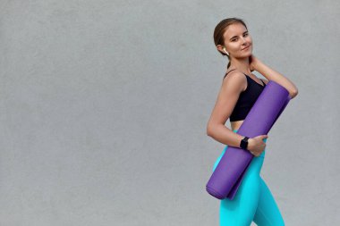 Genç ve sportif bir kadın yoga minderiyle dışarı çıkıyor. Spor kıyafetleri içinde, sağlık ve mutluluk yayıyor, refah ve rahatlığa olan bağlılığını yansıtıyor..