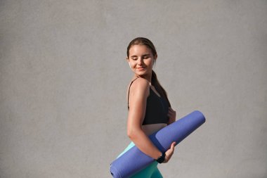 Atletik giyinen genç bir kadın, yoga minderi tutarken gülümserken, sağlıklı bir yaşam tarzı ve zindelik coşkusu sergilerken, tarafsız bir geçmişe sahip, neşeli ifadesini ve aktif ruhunu vurguluyor..