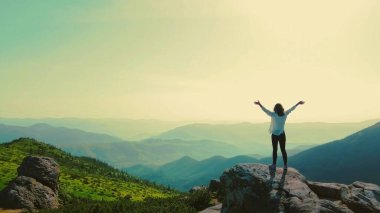 Gün doğumunda bir dağda kollarını kaldırmış bir kadın. Yalnız bir kadın, kolları havada bir dağın tepesinde duruyor, güneşin doğuşunu ve yeşil vadilerin ve uzak dağların geniş manzarasını kutluyor.