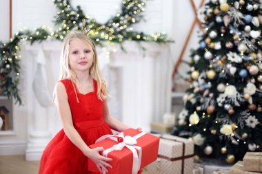 Kırmızı Elbiseli Küçük Kız Noel Hediyesi Tutuyor. Kırmızı elbiseli genç bir kız elinde bir Noel hediyesi, Noel ağacı ve parlak ışıklarla süslenmiş bir odada duruyor..