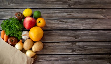 Tahta Masa 'da çeşitli taze ürünler. Domates, patates, havuç, soğan, sarımsak, elma, limon, maydanoz ve kahverengi bir kesekağıdından dökülen portakal gibi çeşitli taze ürünler.