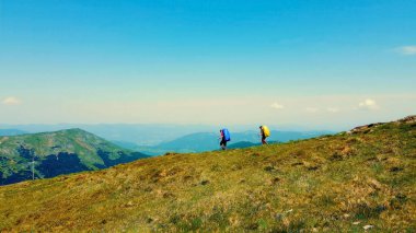 Sırt çantalı yürüyüşçüler dağlık arazide geziniyorlar. Büyük sırt çantalı iki yürüyüşçü, parlak mavi bir gökyüzünün altında manzaralı, çimenli bir dağ sırtından geçiyor.