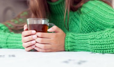 Bir bardak çay tutan ellerin yakın çekimi. Yeşil örgü süveter giyen birinin elinde çay dolu cam bir bardak tutarken yakın plan fotoğrafı.
