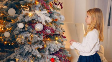 Küçük kız süslü bir Noel ağacına hayran. Genç sarışın bir kız süslerle, ışıklarla ve şenlikli süslemelerle süslenmiş güzel süslenmiş bir Noel ağacına bakıyor.