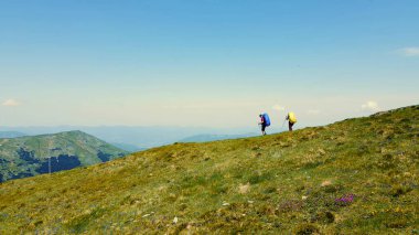 Bir dağ sırtında panoramik manzaralı yürüyüşçüler. Sırt çantalı iki yürüyüşçü bir dağ sırtı boyunca yürüyorlar. Uzaktaki tepelerin ve açık gökyüzünün nefes kesen panoramik görüntüsüyle çevrili.