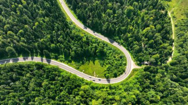 Yoğun yeşil ormanda dolambaçlı dağ yolu. Virajlarda gezinen arabalarla, dağlık bir bölgede, gür ve sık bir ormanın içinden geçen, dolambaçlı bir yolun hava görüntüsü.