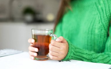 Elinde yeşil kazaklı bir bardak çay tutan kişi. Yeşil örgü süveter giyen birinin elinde çay dolu cam bir bardak tutarken yakın plan fotoğrafı.