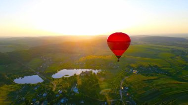 Sıcak Hava Balonu Sunset 'in kırsalında. Güneşin doğuşunda yemyeşil bir kırda süzülen kırmızı bir sıcak hava balonunun nefes kesici hava manzarası..