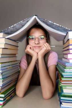 Kitap Yığını Olan Öğrenci. Gözlüklü genç bir kız uzun kitap yığınlarıyla çevrili bir masada oturuyor.