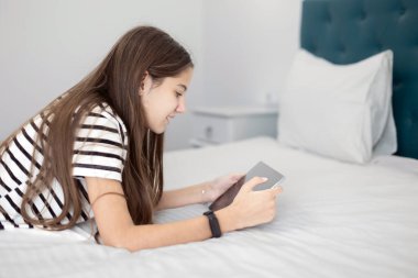 Teenage Girl Using Tablet in a Comfortable Bedroom. A teenage girl with long brown hair lying on a bed while using a tablet clipart