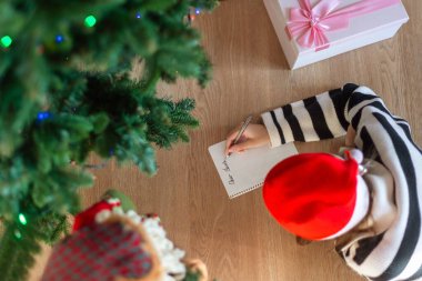 Child Writing Letter to Santa Near Christmas Tree. Overhead view of a child wearing a Santa hat, writing a letter to Santa next to a decorated Christmas tree and gift box clipart