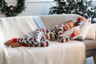 Kanepede şenlikli pijama giyen kız. Noel temalı pijamalı genç bir kız tatil süsleri ve yakındaki ışıklı bir ağaçla rahat bir kanepede uyur.