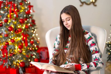 Noel ağacının yanında kitap okuyan bir kız. Güzel dekore edilmiş bir Noel ağacının olduğu bir kitabı okumaya odaklanmış, beyaz bir koltukta oturan neşeli pijamalı genç bir kız.