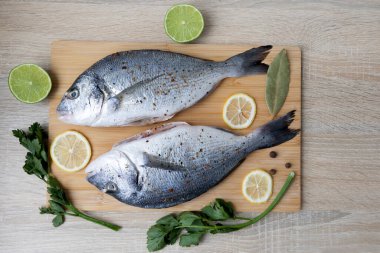 Limonlu taze balık ve kesme tahtasında bitkiler. İki taze balık, limon dilimleri, kireç parçaları, körfez yaprakları, karabiber ve taze bitkilerle çevrili ahşap bir kesim tahtasına yerleştirilir.