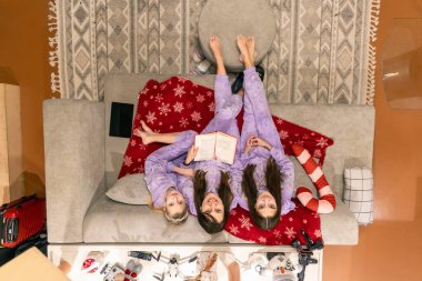Anne ve iki kızı rahat bir kanepede dinleniyorlar. Bir anne ve iki kızının aynı mor pijamaları giyip, kitap okudukları, tatil dekorlarıyla çevrili bir şekilde tepeden tırnağa görüntüsü.
