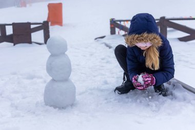 Girl Building Snowman on Winter Day. Young girl wearing winter clothing, shaping snow to build a small snowman outdoors on a snowy day in a scenic setting clipart