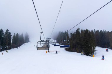 Sisteki Karlı Dağ yamacında kayak kaldırma. Boş kayak kaldırma koltukları karla kaplı bir dağ yamacının üzerinde süzülüyor. Etrafı yemyeşil ağaçlarla çevrili, uzakta kayakçılar ve sis var.