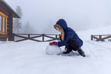 Karlı bir günde kardan adam yapan çocuk. Karda yuvarlanan bir çocuk, sisli bir günde ahşap bir kulübede, arka planda karlı bir yamaçta görünen bulanık figürlerle.