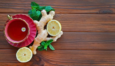 Herbal Tea with Ginger and Lemon Slice. A cup of herbal tea with a chamomile flower, surrounded by fresh ginger, lemon halves, and mint leaves on a wooden table. Top view. Free space for text clipart