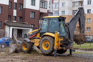 Konut binalarının yakınında kepçe doldurucu çalışıyor. Sarı bir kepçe yükleyici aktif olarak apartman binalarının yakınındaki çamurlu bir alanda kazıyor.