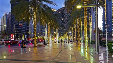 BAE, Dubai 01 Mayıs 2024. Aydınlanmış Palm Trees ile Dubai Caddesi. Modern gökdelenler ve insanlarla çevrili dekoratif ışıklarla kaplı bir Dubai caddesinin akşam manzarası