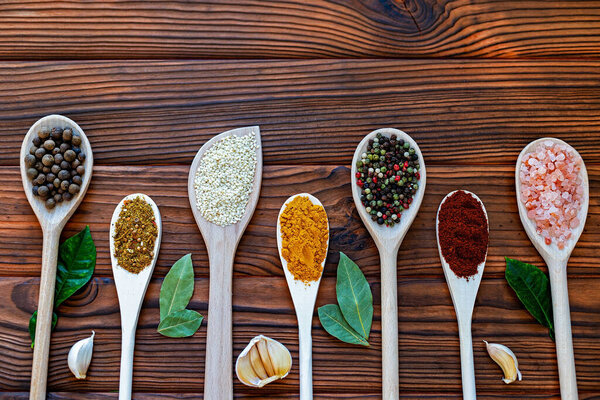 Разнообразные специи на деревянных ложках над деревенским столом. Разнообразие красочных специй, в том числе перца, куркумы, кунжута и гималайской соли, расположенных на деревянных ложках на темном деревянном фоне