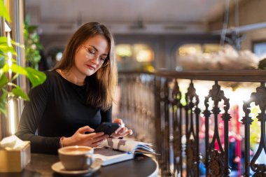 Kafe masasında akıllı telefon okuyan bir kadın, kafede gözlüklü genç bir kadın telefonunda mesajlar okuyor.