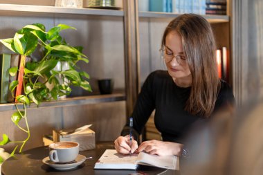 Rahat bir kafede deftere yazan bir kadın. Gözlüklü bir kadın, kafeterya masasındaki bir deftere bir fincan kahve ve yakınlardaki kapalı alan bitkileriyle birlikte yazıyor.