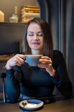 Gözleri kapalı kahvenin tadını çıkaran bir kadın. Gözleri kapalı bir kadın, bir fincan kahveyi elinde tutuyor, bir kafe masasında aromasının tadını çıkarıyor, etrafını kitaplar ve ılık bir ortam sarmış.