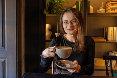 Elinde kahve tutan ve gülümseyen bir kadın. Bardaklı gülümseyen bir kadın kafeterya masasında bir fincan kahve tutuyor, arka planda kitaplar ve bitkiler var.