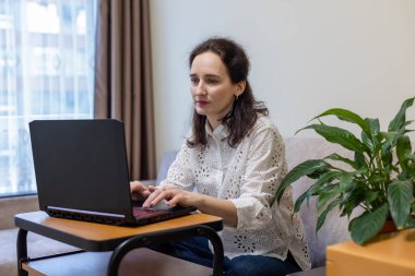 Evde dizüstü bilgisayarla çalışan bir kadın. Beyaz dantelli bluz giyen bir kadın evdeki dizüstü bilgisayarında çalışıyor. Yanında tahta bir masa ve çiçek olan bir kanepede oturuyor..