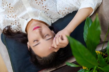 Rahat bir kadın kanepeye uzanmış, gözleri kapalı. Beyaz bluzlu bir kadın kanepeye uzanmış, gözleri kapalı, yastığın üzerinde dinleniyor. Ön planda yeşil bir bitki var..