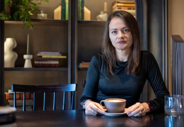 Rahat bir kafede oturan kendine güvenen bir kadın elinde bir fincan kahve, arka planda kitaplık.