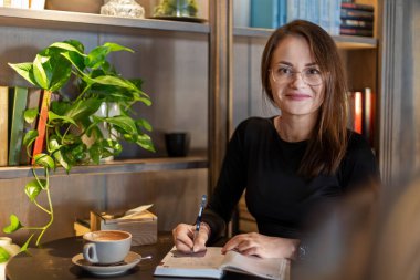 Gülümseyen kadın rahat bir kafede defter, kahve fincanı ve arka planda kitap rafı yazıyor.