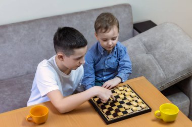 Dama oynayan iki kardeş, beyaz gömlekli büyük kardeş, küçük çocuğa kanepede ders veriyor.