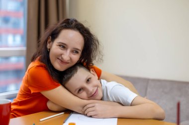 Mutlu Anne Kucaklayan Oğul Evinde. Turuncu gömlekli, sevgi dolu bir anne, sıcak bir evde, masada, gülümseyen oğluna sarılır.