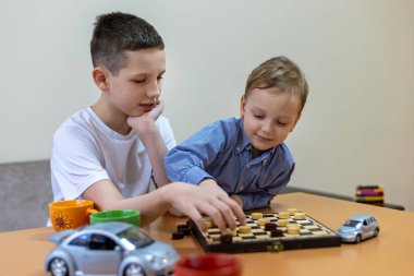 Evde dama oynayan iki çocuk. Masada dama oynayan iki genç çocuk, odaklanmış ve meşgul, oyuncak arabalar ve renkli kupalarla çevrili..